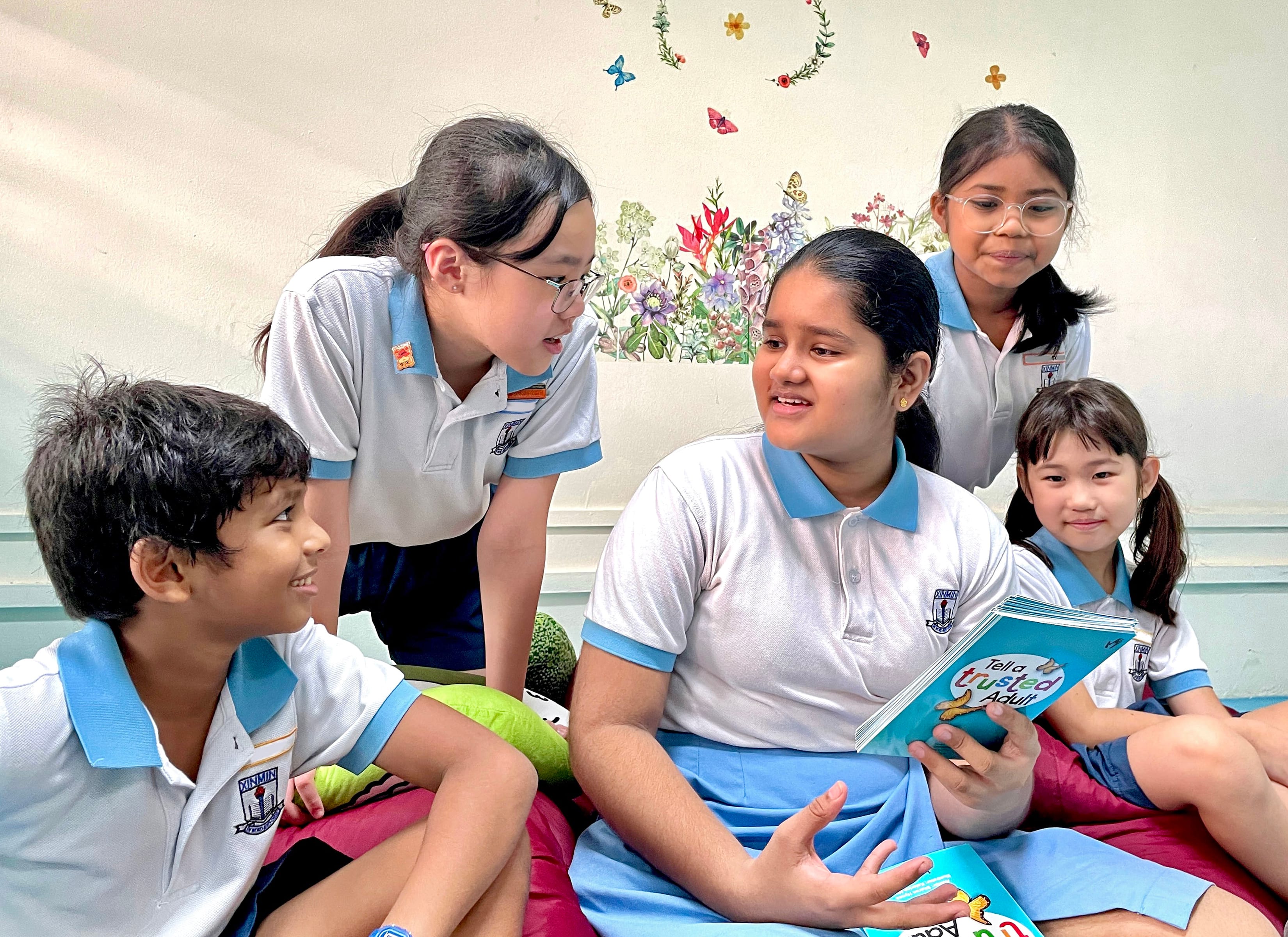 Five children in school uniforms sit together as one girl reads aloud a book titled Tell a Trusted Adult.